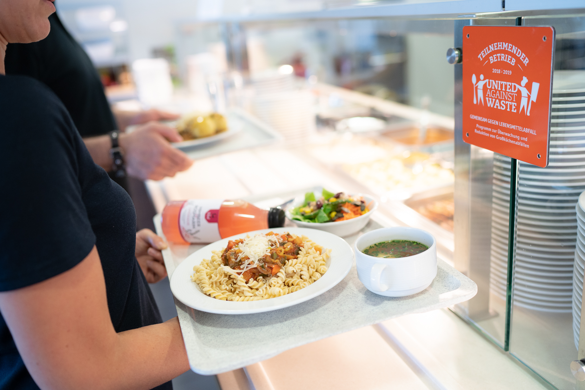 Person hält gefülltes Tablett in Kantine und schaut auf eine Plakette von United Against Waste