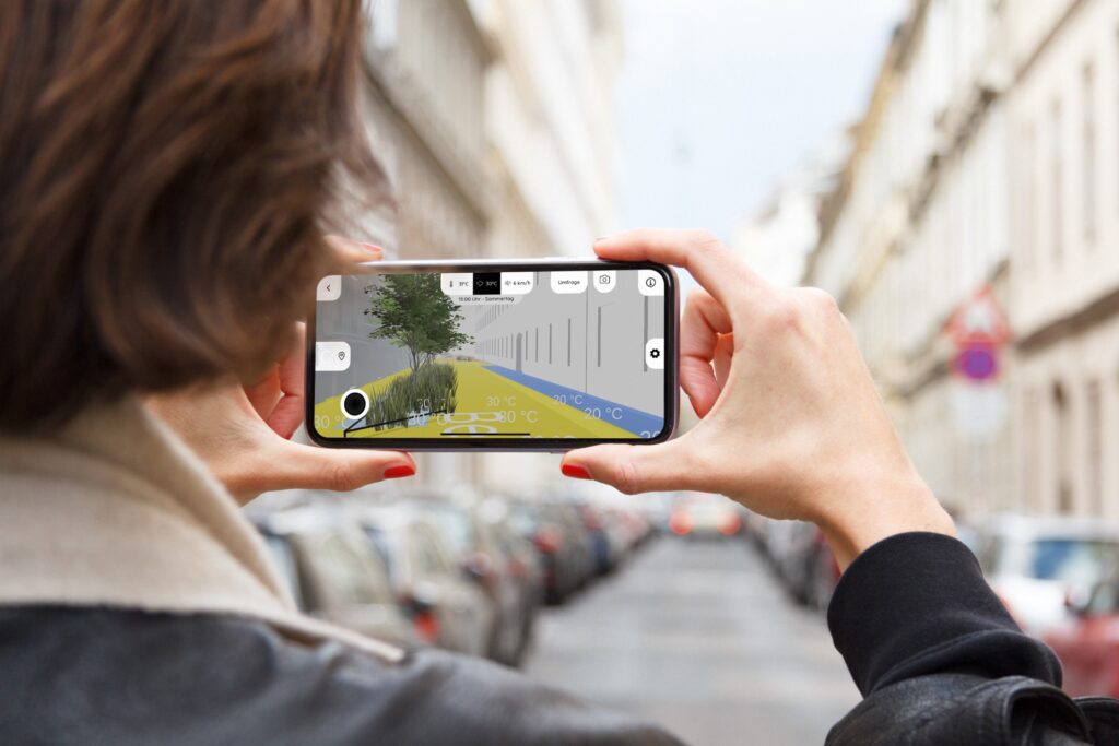 Person hält Smartphone vor einer grauen Straße und sieht eine Grafie, wie die Straße begrünt aussehen würde inklusive Anzeige der Temperatur auf der Straße